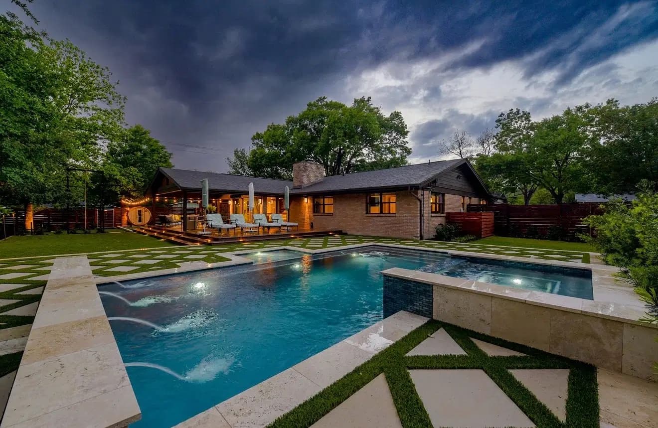 Geometric backyard pool and raised spa beside a brick home under a cloudy evening sky